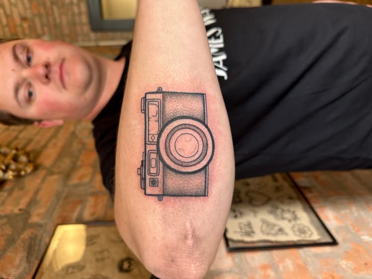 Black and gray camera tattoo on a person's forearm with fine line details
