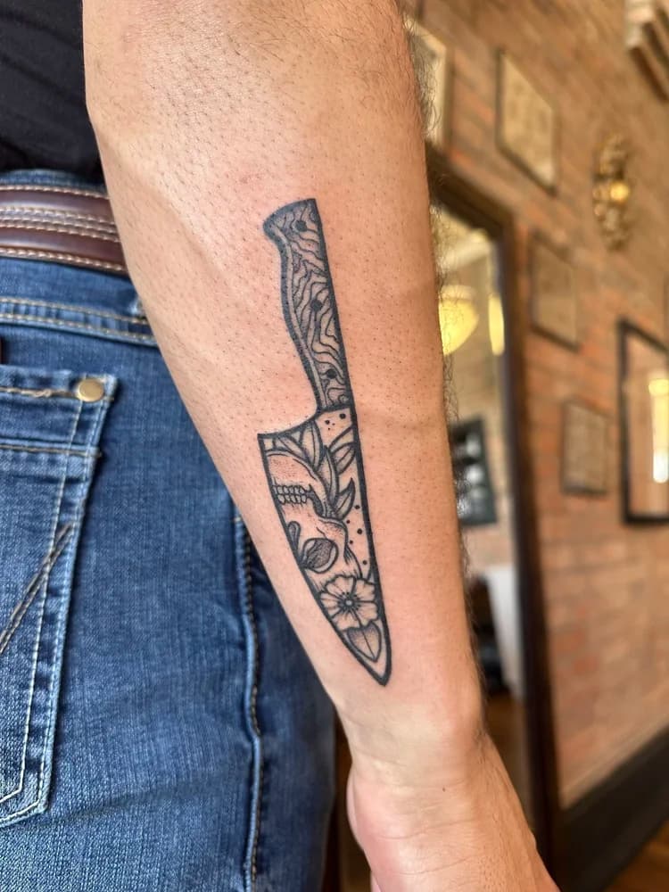 Black and gray decorative knife tattoo on forearm with ornamental floral patterns